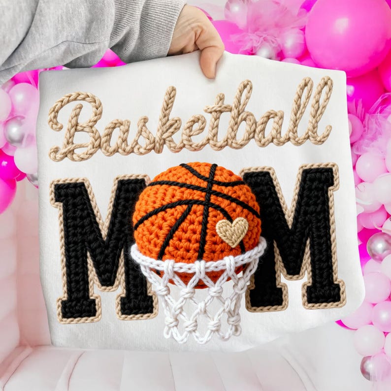 May include: A white decorative pillow with the words "Basketball Mom" embroidered in a rope-like material. The "MOM" letters are black with a white border. An orange basketball with a heart is in the center, above a white net.