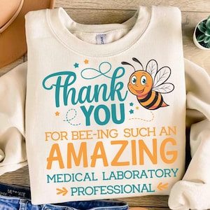 Puede incluir: Una sudadera beige con un gráfico de abeja y el texto "Gracias por ser un(a) profesional de laboratorio médico increíble".