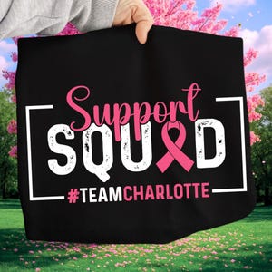 Può includere: Borsa tote nera con le parole "Support Squad" in corsivo rosa e lettere bianche. Un nastro rosa è incorporato nel design. Il testo "#TEAMCHARLOTTE" è sotto in bianco.