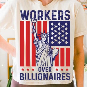 Könnte beinhalten: Cremefarbenes T-Shirt mit einer Grafik, die die Freiheitsstatue mit einem Schraubenschlüssel vor einem rot-weiß-blauen amerikanischen Flaggen-Design zeigt. Der Text lautet "WORKERS OVER BILLIONAIRES."