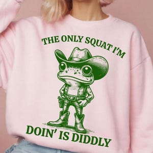 Może przedstawiać: R&oacute;żowa bluza z zieloną kresk&oacute;wkową żabą w kowbojskim kapeluszu i tekstem "The only squat I'm doin' is diddly."