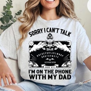 May include: Light gray t-shirt with the text "SORRY I CAN'T TALK I'M ON THE PHONE WITH MY DAD". The shirt features a black Ouija board design with bats, skulls, and the alphabet.