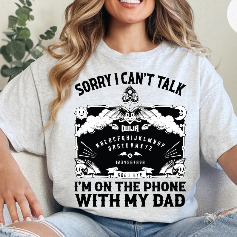 May include: Light gray t-shirt with the text "SORRY I CAN'T TALK I'M ON THE PHONE WITH MY DAD". The shirt features a black Ouija board design with bats, skulls, and the alphabet.