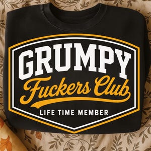 May include: Black sweatshirt with a graphic that reads "GRUMPY Fuckers Club" in white and gold lettering. The text is enclosed in a white and gold outlined shape. Below the text is the phrase "LIFE TIME MEMBER".