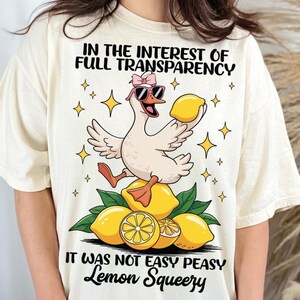 May include: Cream-colored t-shirt with a whimsical design. It features a cartoon goose wearing sunglasses and a bow, holding a lemon. The text reads "IN THE INTEREST OF FULL TRANSPARENCY" and "IT WAS NOT EASY PEASY Lemon Squeezy".