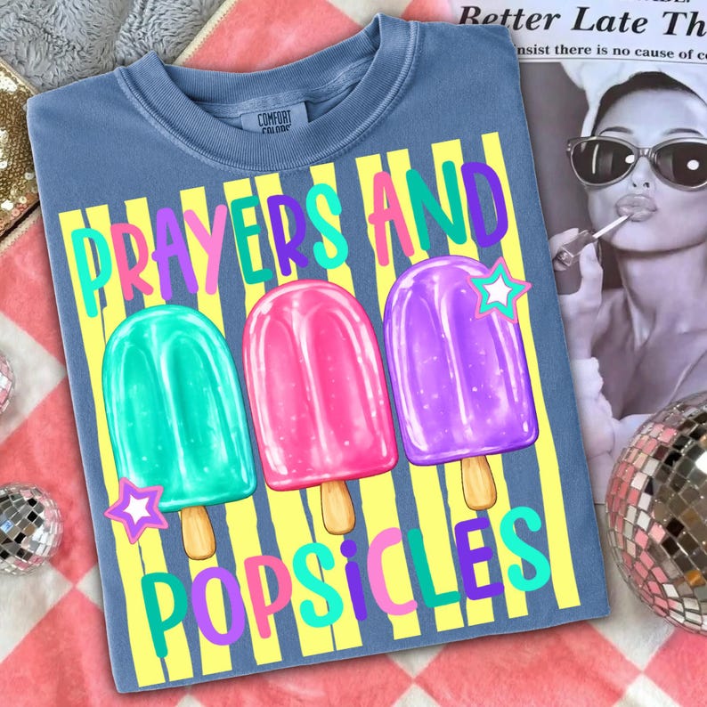 K&ouml;nnte beinhalten: Blaues T-Shirt mit einer Grafik von drei bunten Eis am Stiel auf gelben und wei&szlig;en Streifen. Der Text "Prayers and Popsicles" ist in leuchtenden Farben gehalten. Das Shirt hat ein Comfort Colors-Etikett.