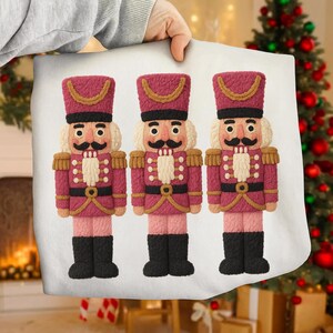 May include: A white pillow featuring three decorative nutcracker soldiers. Each nutcracker has a burgundy hat and uniform with gold accents, a black mustache, and black boots. The pillow is held by a person.