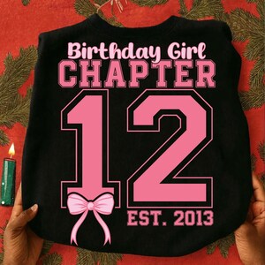 Può includere: Felpa nera con la scritta "Birthday Girl Chapter 12" in rosa. Il numero 12 è grande e rosa, con un fiocco rosa in basso. Il testo "EST. 2013" è sotto il numero.