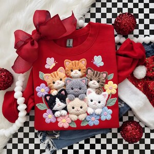 Pu&ograve; includere: Una felpa rossa con un disegno ricamato di sette gattini circondati da fiori e farfalle. La felpa ha un girocollo e maniche lunghe. Sono visibili anche un grande fiocco rosso e un cappello da Babbo Natale.