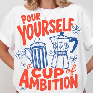 May include: A white t-shirt featuring a graphic print with the red text "POUR YOURSELF a CUP of AMBITION". The design includes a blue striped coffee mug and a blue moka pot, surrounded by small blue flower illustrations on the front of the shirt.