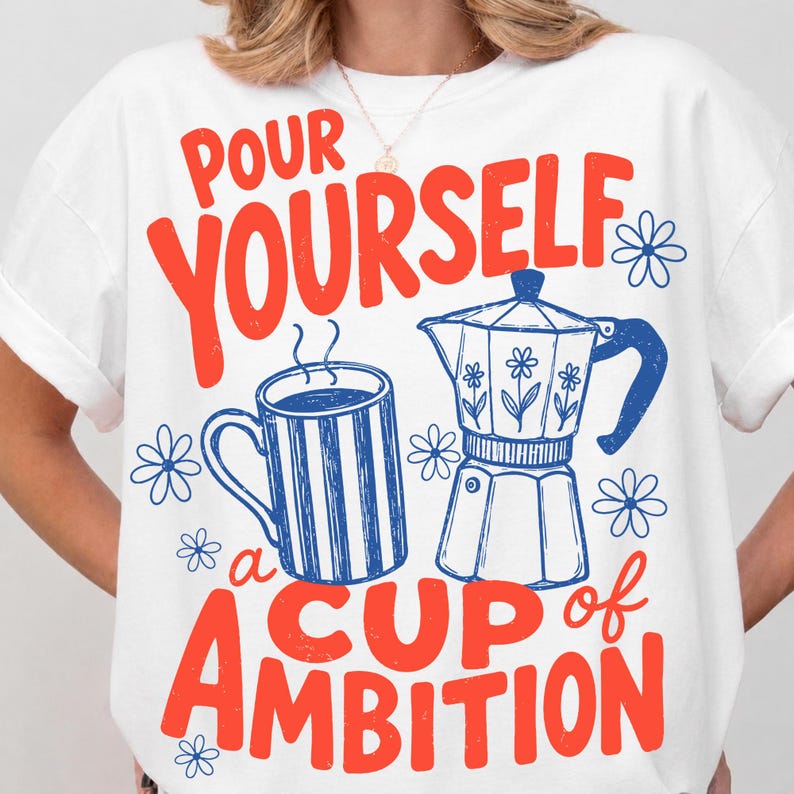 May include: A white t-shirt featuring a graphic print with the red text "POUR YOURSELF a CUP of AMBITION". The design includes a blue striped coffee mug and a blue moka pot, surrounded by small blue flower illustrations on the front of the shirt.