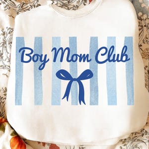 Op de afbeelding: Witte sweatshirt met blauwe verticale strepen en de tekst "Boy Mom Club" in een scriptlettertype. Een blauwe strik-afbeelding is gecentreerd onder de tekst. De sweatshirt is een casual, comfortabele stijl.