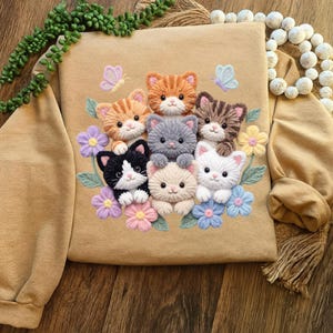 Pu&ograve; includere: Felpa beige con un ricamo raffigurante sette gattini colorati circondati da fiori e farfalle. I gattini sono di vari colori, tra cui arancione, grigio e bianco. I fiori sono in tonalit&agrave; di viola, rosa e blu.