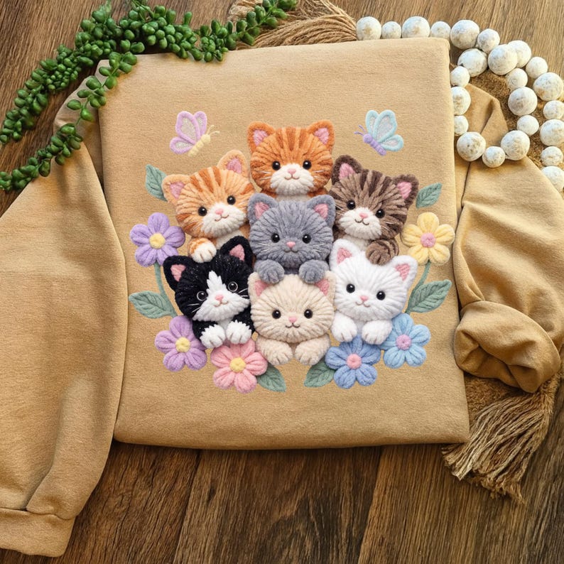 Pu&ograve; includere: Felpa beige con un ricamo raffigurante sette gattini colorati circondati da fiori e farfalle. I gattini sono di vari colori, tra cui arancione, grigio e bianco. I fiori sono in tonalit&agrave; di viola, rosa e blu.