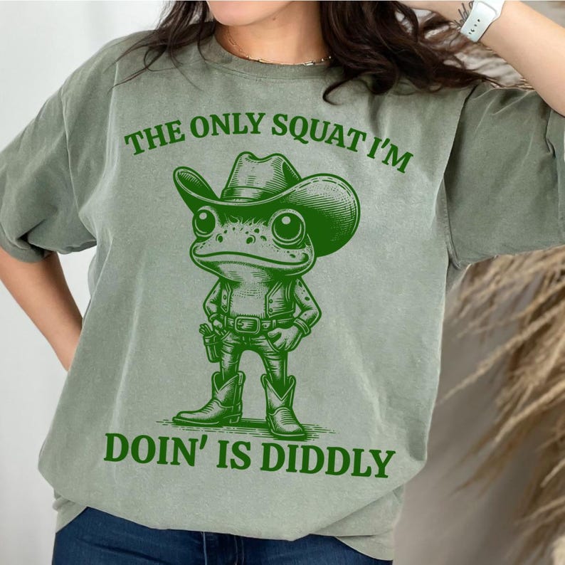 Może przedstawiać: Zielony T-shirt z kresk&oacute;wkową żabą w kowbojskim kapeluszu i trzymającą pistolet. Tekst na koszulce brzmi "The only squat I'm doin' is diddly."