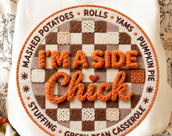 May include: Cream-coloured sweatshirt with an embroidered circular design. The design features the text "I'M A SIDE Chick" in orange yarn, a brown and white checkered pattern, and the words "Mashed Potatoes", "Stuffing", "Rolls", "Yams", "Pumpkin Pie", and "Green Bean Casserole".