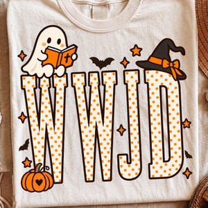 May include: A cream-colored t-shirt featuring the letters WWJD in orange polka dots. The design includes a white ghost reading an orange book, a black witch hat with an orange ribbon, small black bats, and orange stars, plus an orange pumpkin.