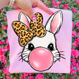May include: A light pink sweatshirt featuring a cartoon bunny with a large pink bubble gum bubble. The bunny has a white face, black eyes, and a leopard print bow. The background is a vibrant display of pink flowers.