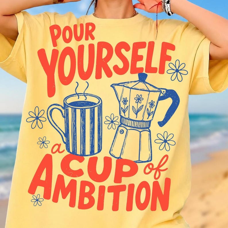 May include: A pale yellow t-shirt featuring a graphic print in red and blue. The design includes a striped coffee mug, a moka pot, and small flower icons. The text reads: POUR YOURSELF a CUP of AMBITION in bold, stylized red lettering.