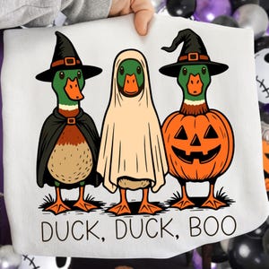 May include: A white Halloween-themed graphic featuring three ducks dressed in costumes. One duck is a witch, one is a ghost, and one is a jack-o'-lantern. The text "DUCK, DUCK, BOO" is printed at the bottom of the image.