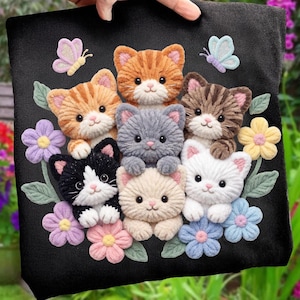 Puede incluir: Bolso tote negro con un diseño de siete gatitos de peluche de varios colores, rodeados de flores y mariposas. Los gatitos son naranjas, grises, blancos y negros y blancos. El bolso es sostenido por una mano.