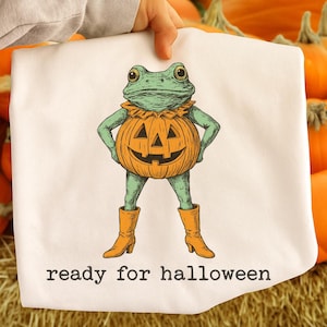 May include: White Halloween-themed fabric featuring a green frog with a pumpkin body and orange boots. The frog has its hands on its hips and is ready for Halloween. The text "ready for halloween" is printed at the bottom.