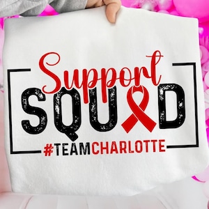 May include: White tote bag with the words "Support Squad" in red script and black block letters. A red ribbon symbol is incorporated into the design. The text "#TeamCharlotte" is below the main text.