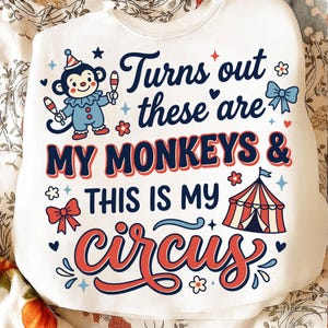 May include: White sweatshirt with a colorful graphic. The design features a cartoon monkey, circus tent, and the text "Turns out these are MY MONKEYS & THIS IS MY circus" in blue and red lettering. Decorative stars and flowers surround the text.