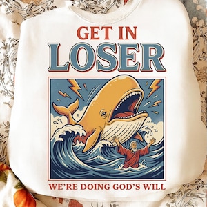 May include: White sweatshirt with a graphic design featuring a yellow whale, blue waves, and a man in red robes. The text "GET IN LOSER" is above the image, and "WE'RE DOING GOD'S WILL" is below.