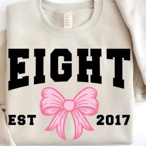 Pode incluir: Uma camisola de gola redonda bege com a palavra EIGHT em letras pretas grandes no peito. Abaixo do texto encontra-se uma ilustração gráfica de um laço cor-de-rosa claro. O texto EST 2017 está impresso a preto em ambos os lados do laço.