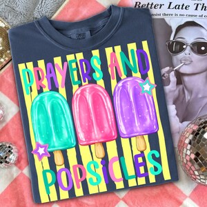 K&ouml;nnte beinhalten: Dunkelblaues T-Shirt mit den Worten "PRAYERS AND POPSICLES" in farbenfrohem Text. Das Shirt zeigt drei Eis am Stiel-Illustrationen in Gr&uuml;n, Pink und Lila, jede mit einem Stern. Das Shirt liegt auf einer rot-wei&szlig; karierten Oberfl&auml;che.