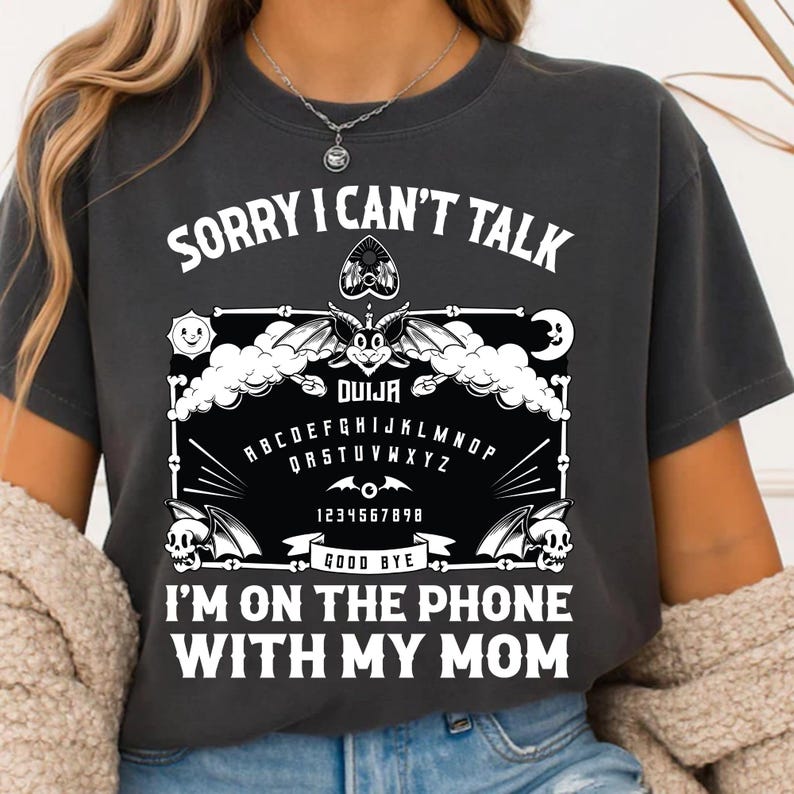 May include: Dark gray t-shirt with white text and a Ouija board design. The text reads "SORRY I CAN'T TALK" at the top and "I'M ON THE PHONE WITH MY MOM" at the bottom. The Ouija board graphic includes the alphabet, numbers, and the word "DUIJA".