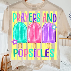 K&ouml;nnte beinhalten: Beiges T-Shirt mit dem Schriftzug "Prayers and Popsicles" in bunten Farben. Das Design zeigt drei Eis am Stiel-Illustrationen in T&uuml;rkis, Pink und Lila, jeweils mit einem Stern. Das Shirt hat einen Rundhalsausschnitt.