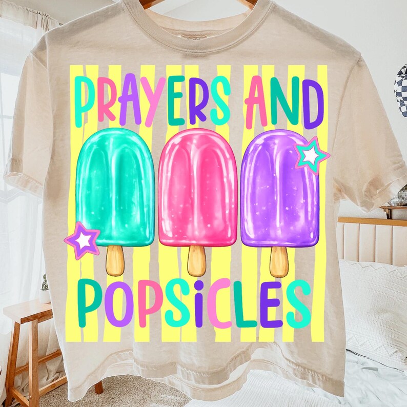 K&ouml;nnte beinhalten: Beiges T-Shirt mit dem Schriftzug "Prayers and Popsicles" in bunten Farben. Das Design zeigt drei Eis am Stiel-Illustrationen in T&uuml;rkis, Pink und Lila, jeweils mit einem Stern. Das Shirt hat einen Rundhalsausschnitt.