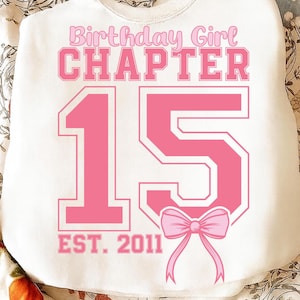 May include: White sweatshirt with pink text that reads "Birthday Girl CHAPTER 15 EST. 2011" with a pink bow graphic. The number 15 is large and prominent. The sweatshirt is a birthday-themed apparel item.