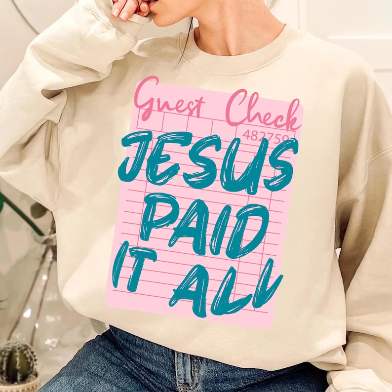 May include: A beige sweatshirt featuring a pink graphic resembling a guest check. The graphic reads "Guest Check" in pink, and "JESUS PAID IT ALL" in teal. The sweatshirt is a casual style.