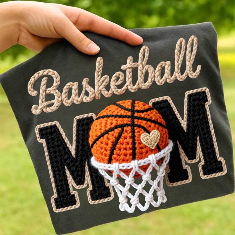 May include: Dark green t-shirt with the words "Basketball Mom" in a rope-like font. The design includes a crocheted basketball, net, and the letters "M" in black with a cream border. A small crocheted heart is on the basketball.