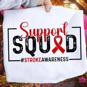 May include: White fabric with the words "Support Squad" in red and black, with a red ribbon symbol. The text "#StrokeAwareness" is below. The fabric is held up by a person.