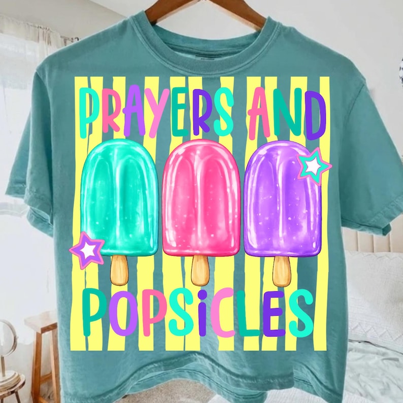 K&ouml;nnte beinhalten: T&uuml;rkisfarbenes T-Shirt mit dem Schriftzug "Prayers and Popsicles" in bunten Farben. Das Design zeigt drei Eis am Stiel-Illustrationen in Gr&uuml;n, Pink und Lila, jeweils mit einem Stern. Gelbe vertikale Streifen bilden den Hintergrund.