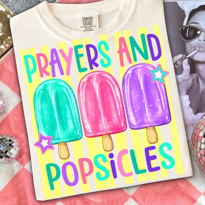 K&ouml;nnte beinhalten: Cremefarbenes T-Shirt mit den Worten "Prayers and Popsicles" in farbenfrohem Text. Das Design zeigt drei Eis am Stiel-Illustrationen in T&uuml;rkis, Rosa und Lila, jeweils mit einem Stern-Akzent. Das Shirt liegt auf einer rosa-wei&szlig; karierten Oberfl&auml;che.