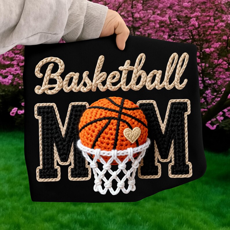 May include: Black decorative pillow with the words "Basketball Mom" in a rope-like font. The design includes a crocheted basketball, net, and a heart. The pillow is held by a person.