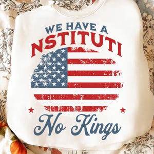 May include: White sweatshirt with the text "WE HAVE A NSTITI No Kings" in red and blue. The design features a distressed American flag graphic in a circular shape, with red stars and stripes.