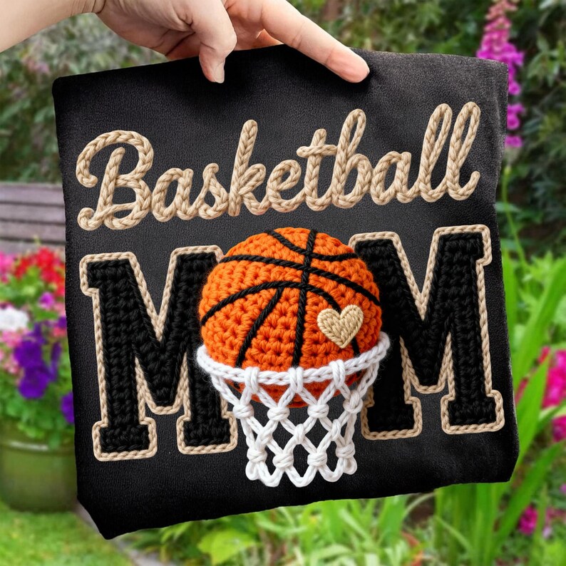 May include: Black fabric with the words "Basketball Mom" in beige rope-style lettering. A crocheted basketball with a heart is centered between the letters "M" and "M". The basketball is orange and black, with a white net.