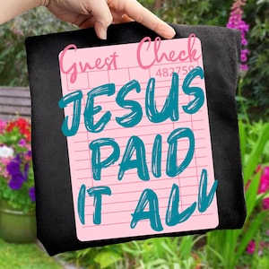 May include: A black tote bag with a pink guest check design. The guest check has the words "Guest Check" in pink script, and the words "JESUS PAID IT ALL" in teal script.
