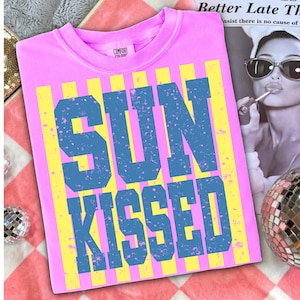 以下が含まれることがあります： 青い「SUN KISSED」の文字がプリントされたピンク色のクルーネックのスウェットシャツ。文字は黄色の縦縞模様の上に配置されています。シャツにはComfort Colorsのタグが付いています。