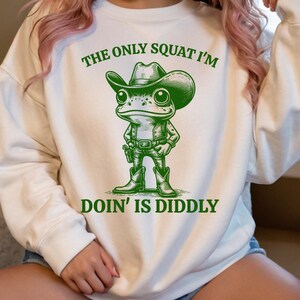 Może przedstawiać: Biała bluza z zieloną żabą w kowbojskim kapeluszu i butach. Tekst na bluzie brzmi "The only squat I'm doin' is diddly."