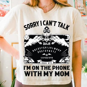 May include: Cream-colored t-shirt with a black Ouija board graphic. The graphic includes the text "SORRY I CAN'T TALK", "DUIJA", the alphabet, numbers, and "GOOD BYE". The bottom of the shirt reads "I'M ON THE PHONE WITH MY MOM".