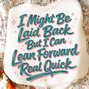 以下が含まれることがあります： 白のスウェットシャツ。テキスト「I Might Be Laid Back But I Can Lean Forward Real Quick」は、ティールとピンクの文字で書かれています。デザインには星のアクセントが含まれています。スウェットシャツはおそらく柔らかく快適な素材で作られています。