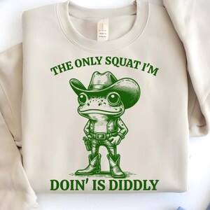 Może przedstawiać: Beżowa bluza z zieloną żabą w kowbojskim kapeluszu i napisem "The only squat I'm doin' is diddly."