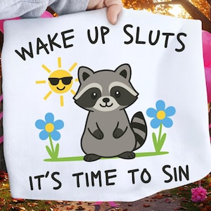 Peut inclure: Tissu blanc avec le texte "WAKE UP SLUTS IT'S TIME TO SIN" en noir. Un raton laveur de dessin animé avec une queue rayée est centré, avec un soleil jaune portant des lunettes de soleil et des fleurs bleues de chaque côté.